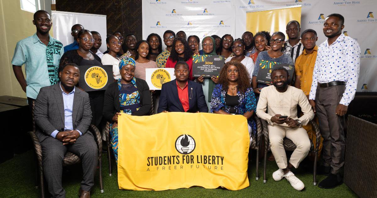 A diverse group of young African men and women, smiling and engaged, pose for a group photo indoors at what appears to be a conference or training event. Many are holding certificates or placards with text related to AfCFTA and economic empowerment. In the foreground, a large yellow banner with "STUDENTS FOR LIBERTY A FREER FUTURE" is displayed. Several individuals are seated in chairs in the front row, while others stand behind them. The background shows hints of a hotel setting with "Airport View Hotel" visible on some signs.