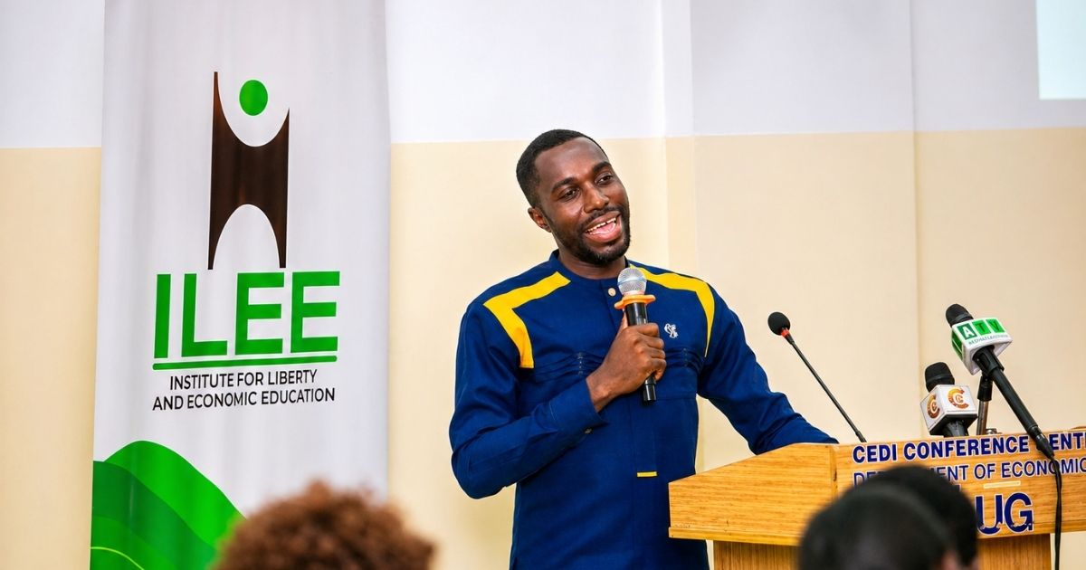 A man speaking at a podium during a conference, holding a microphone, with an ILEE banner in the background.