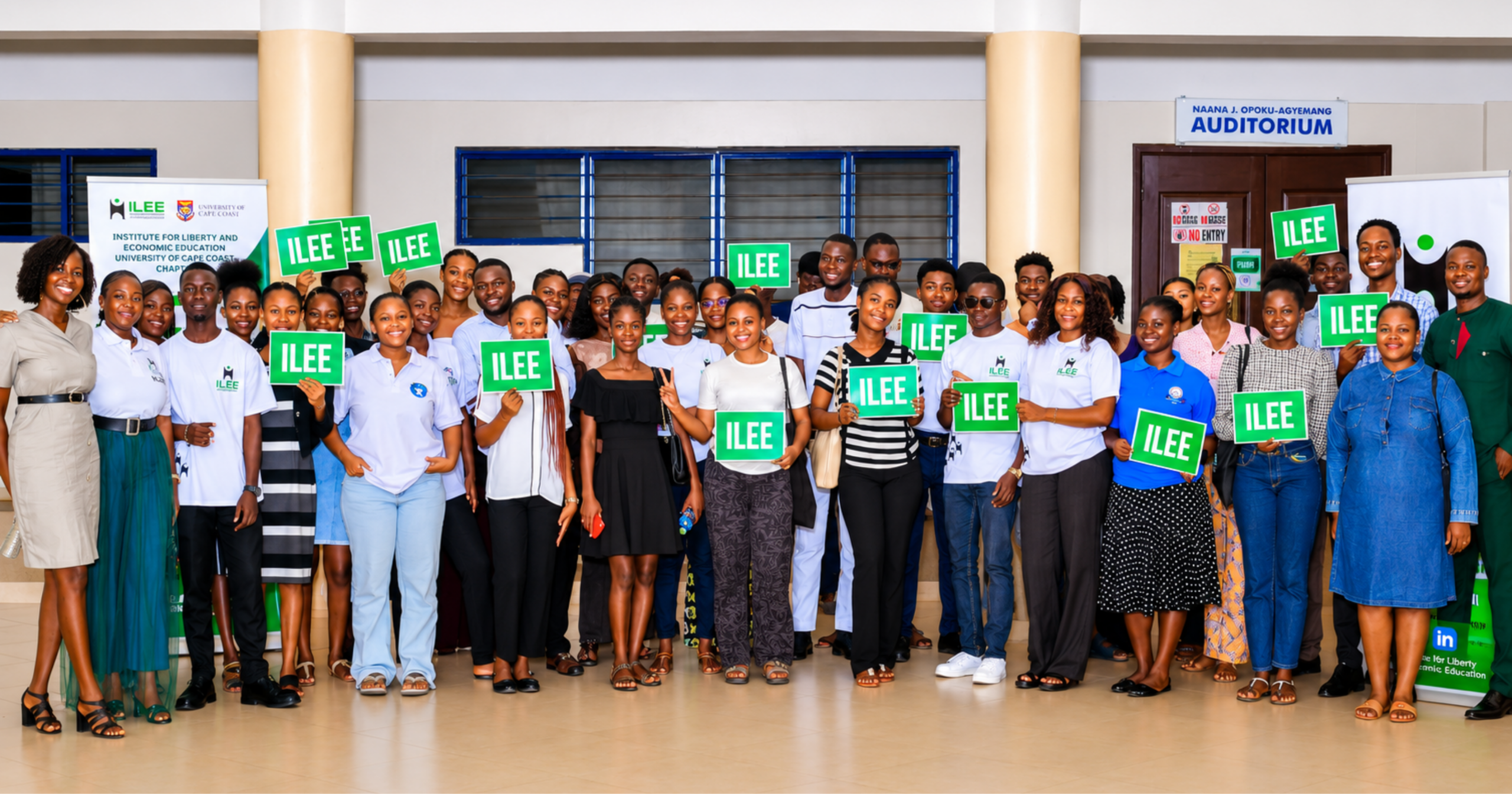 Group photo of ILEE participants and organizers holding ILEE signs at the University of Cape Coast auditorium after an entrepreneurship and economic education event.